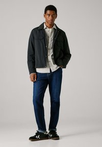 Chaqueta negra con dos bolsillos en el pecho, camisa a cuadros blancos debajo, jeans de mezclilla azules y zapatillas negras con detalles blancos.