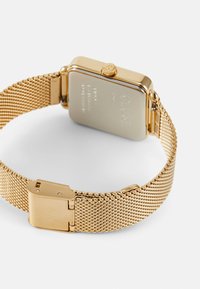 Montre en maille dorée avec un boîtier rectangulaire et une couronne. L'arrière présente des détails gravés, et le fermoir comprend un mécanisme de fermeture sécurisé.