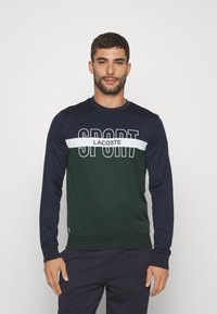 Sudadera de manga larga con un diseño en bloques de colores en verde oscuro y azul marino, con el texto "SPORT" en blanco y la marca "LACOSTE" en el pecho.