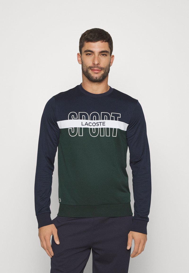 Sudadera de manga larga con un diseño en bloques de colores en verde oscuro y azul marino, con el texto "SPORT" en blanco y la marca "LACOSTE" en el pecho.