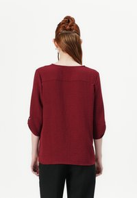 Blusa bordeaux in tessuto texturizzato con scollatura rotonda e maniche a tre quarti. Sfiancata leggermente, con retro semplice senza decorazioni.