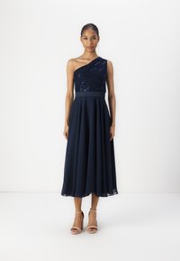 Robe bleu marine à une épaule avec un corsage paillet�é et une jupe fluide en couches. Portée avec des talons à lanières de couleur claire.