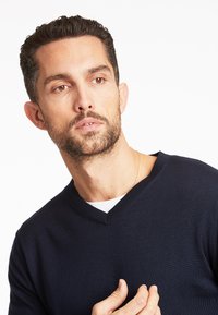 Maglione blu navy a V in tessuto lavorato a maglia; texture sottile; indossato sopra una camicia bianca; il modello ha i capelli corti, mostrando i lineamenti del viso.
