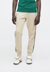 Μπεζ φόρμα με μαύρες ρίγες στο πλάι, κατασκευασμένη από απαλό υλικό, στενή εφαρμογή, συνδυασμένη με λευκά αθλητικά παπούτσια με μαύρες ρίγες και υφασμάτινες λεπτομέρειες.