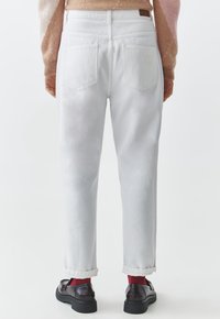 Jeans in denim bianco con una vestibilità rilassata, cuciture visibili e due tasche posteriori. Le cuffie arrotolate rivelano calzini rossi e scarpe nere.