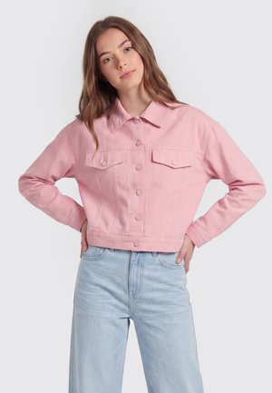 Jonge vrouw die een cropped roze jasje met knopen en lichtblauwe hoge taille jeans draagt, staand met handen in de zij tegen een effen achtergrond.