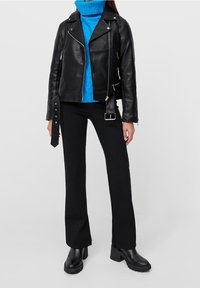 Nainen, jolla on päällään musta nahkatakki kirkkaan sinisen poolopaidan, mustien levenevien housujen ja mustien paksupohjaisten saappaiden kanssa, seisoo valkoisella lattialla.