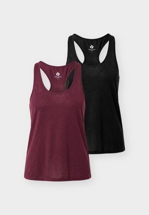 Nuori henkilö seisoo päällään hihaton burgundinpunainen toppi, mustat leggingsit, valkoiset tennarit ja vaaleanharmaat sukat yksiväristä taustaa vasten.