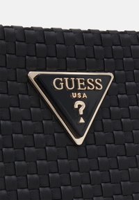 Surface en cuir tressé noir avec un logo triangulaire Guess USA noir et or, comportant un point d'interrogation au centre.