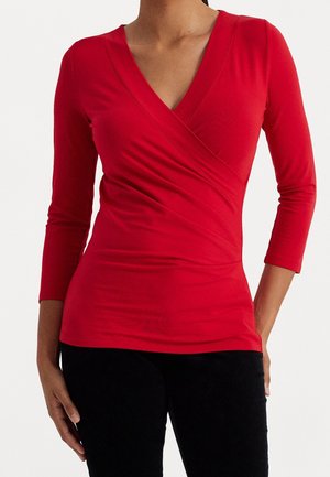 Vrouw draagt een aansluitende, rode wikkelstijl top met lange mouwen en een diep V-hals, gecombineerd met een zwarte broek, staand met één hand langs haar zijde.