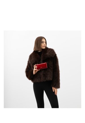 Vrouw in een donkergrijze bontjas en zwarte broek houdt een rode portemonnee vast, ze kijkt naar rechts tegen een effen witte achtergrond.