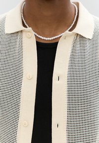 Beżowy sweter z dzianiny o wzorze siateczkowym, zapinany na guziki, z kołnierzykiem. Nosi się go na czarnej, prążkowanej koszulce, w towarzystwie perłowej naszyjnika.