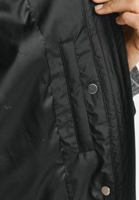 Zwarte gewatteerde jacks binnenkant met een gladde textuur, een zijzak en een drukknoopdetail, met subtiele branding overal.