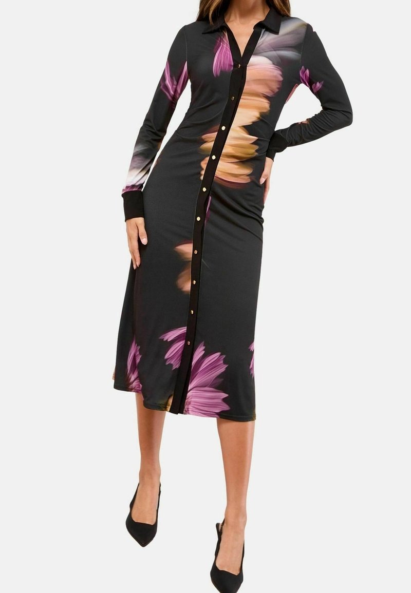Femme portant une robe midi noire à manches longues avec de grands imprimés floraux roses et orange, et des talons hauts noirs.