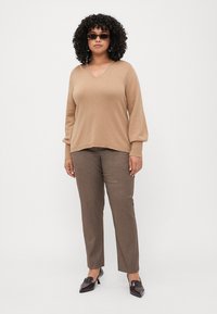 Pull beige en V à manches longues, associé à un pantalon marron ajusté et des chaussures à bout pointu noires, accessoirisé avec des lunettes de soleil noires.