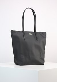 Sac fourre-tout noir avec poignées en cuir, fermeture éclair, et petit logo crocodile vert sur le devant, posé sur une surface blanche.