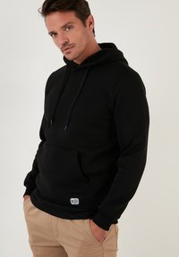 Sudadera negra hecha de tela suave, con un bolsillo frontal, capucha con cordón y puños acanalados. Diseño simple con un detalle de marca mínimo.
