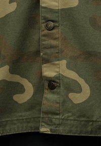 Κοντινή εικόνα από το μπροστινό μέρος πουκάμισου με ύφασμα παραλλαγής πράσινου και καφέ χρώματος και δύο ορειχάλκινα κουμπιά με την ένδειξη «Gardening Wear Co.»