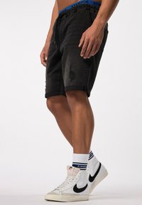 Shorts de mezclilla negra con un patrón desgastado, cinturilla elástica con una marca azul, combinados con zapatillas blancas de caña alta y calcetines con estampado.