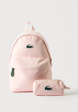 Sac à dos rose clair et trousse assortie avec logos crocodile verts sur fond blanc uni.