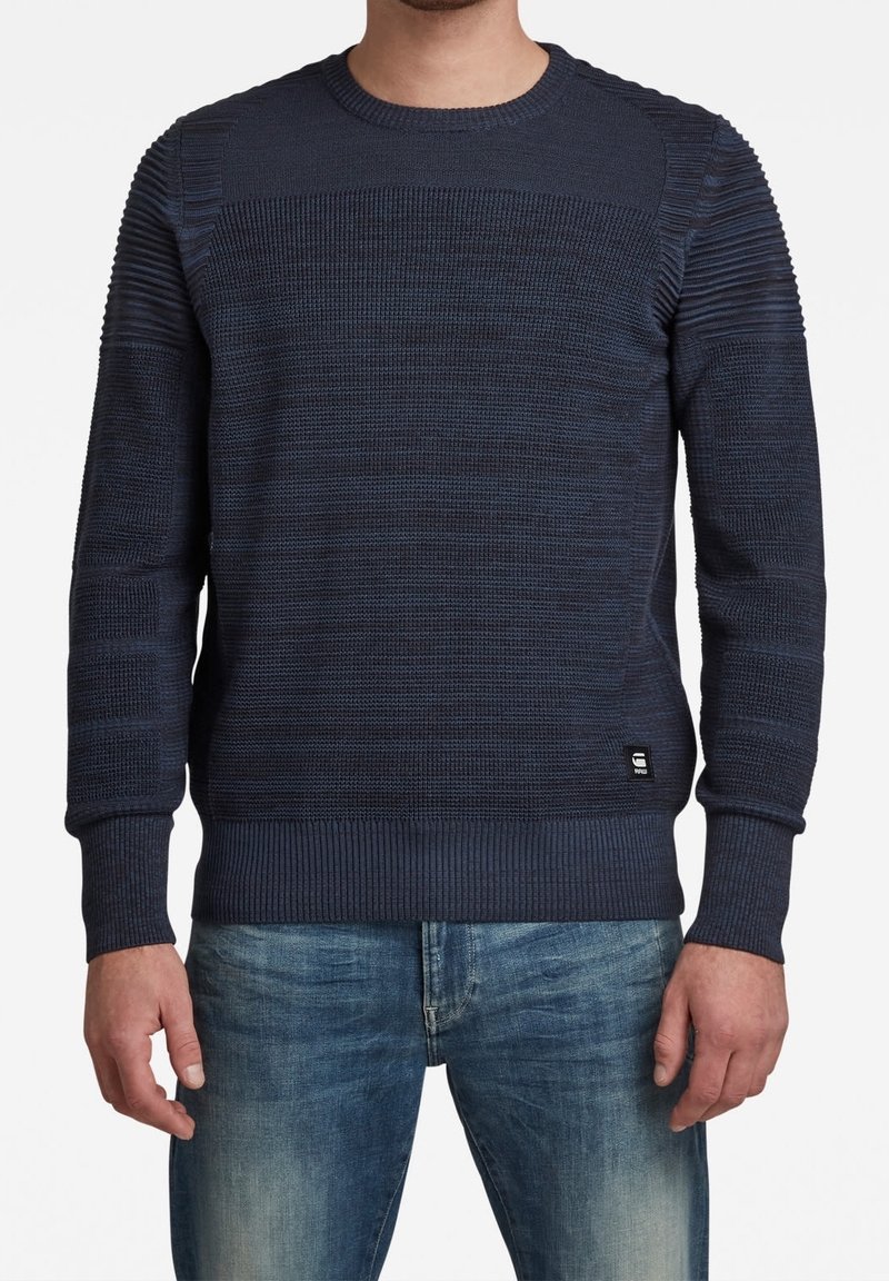 Homme portant un pull en maille texturée bleu foncé avec poignets et ourlet côtelés, associé à un jean bleu délavé, debout devant un fond blanc.