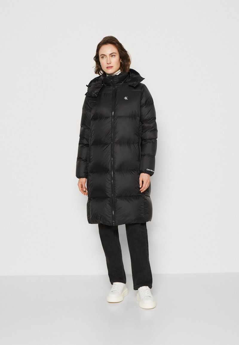 Black puffer coat featuring a zipper front, hood, and logo accents. The coat has a long, oversized fit and is made of smooth, shiny fabric.