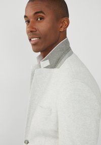 Hellgraue Jacke mit hohem Kragen, die eine weiche Textur und einen kontrastierenden grauen Kragen aufweist. Sie verfügt über eine einzelne Tasche und Frontknöpfe.