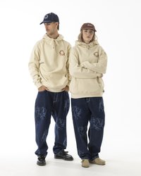 Beige hettegensere med snøring og grafiske detaljer, kombinert med løse blå denimjeans med et mønster. Fottøy inkluderer svarte sko og beige mokkasiner.