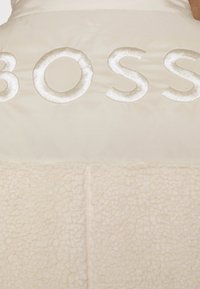 Beige pufferjacka med en plyschshearling-nedre del. Har en stor, broderad "BOSS"-logga i en matchande ljus färg på den övre delen.