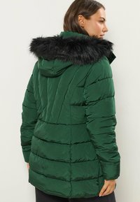 Mörkgrön puffjacka med en svart fuskpälsfodrad huva, kviltat mönster och ribbade ärmslut. Har fickor och en något förlängd bak.