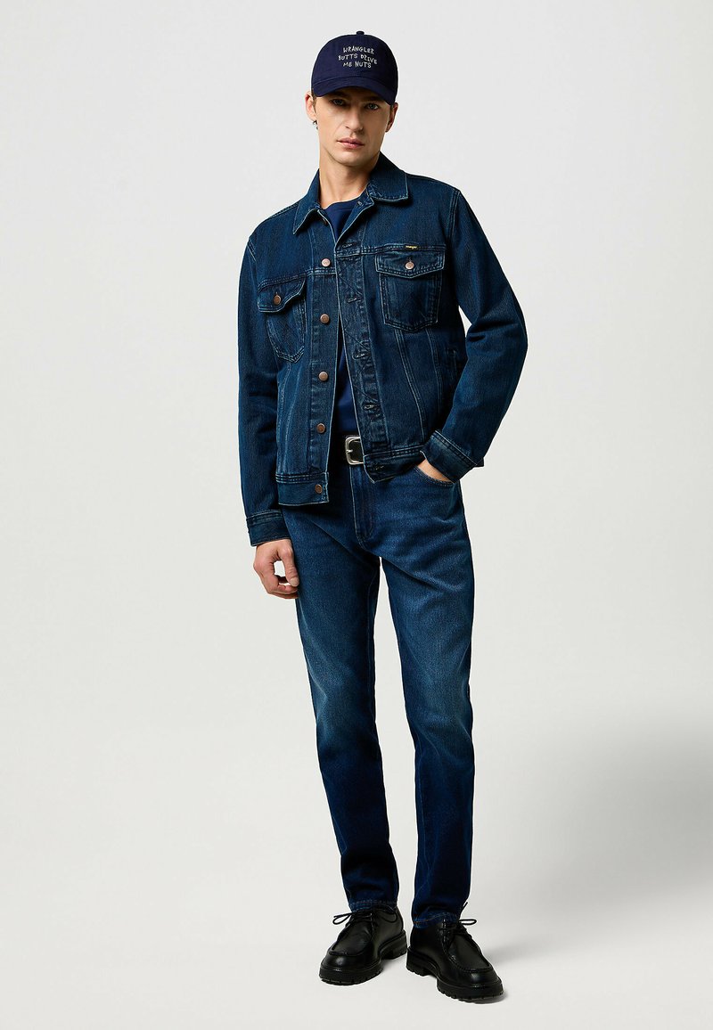 Donkerblauwe spijkerjas met borstzakken, gecombineerd met donkerblauwe jeans. Zwarte schoenen en een marineblauwe pet met tekst aan de voorkant.