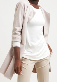 Manteau beige clair, longueur genou, manches longues, revers retroussés, devant ouvert, texture douce. Associé à un haut blanc ajusté et un pantalon beige.