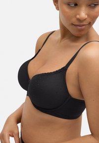 Sostén acolchado negro con tejido texturizado que presenta pequeños puntos. Tirantes ajustables y detalle de encaje a lo largo del borde superior.