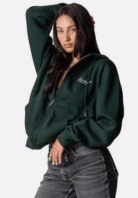Dunkelgrüner Reißverschluss-Hoodie mit Kängurutasche, Kordelzugkapuze und besticktem Logo auf der linken Seite. Übergrößenschnitt mit gerippten Bündchen.