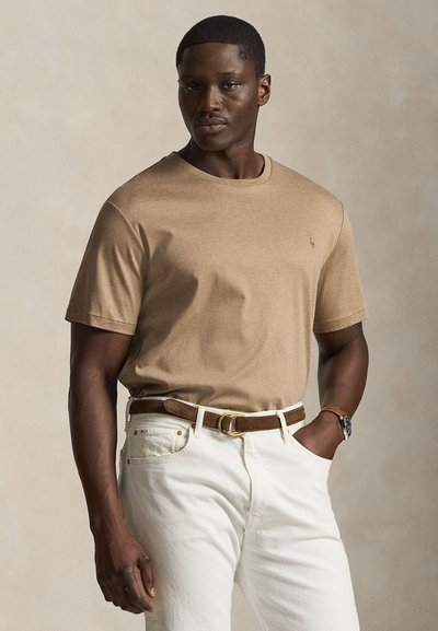 Hombre que lleva una camiseta beige de manga corta, pantalones blancos, cinturón marrón y reloj, de pie con una mano en el bolsillo contra un fondo neutro.