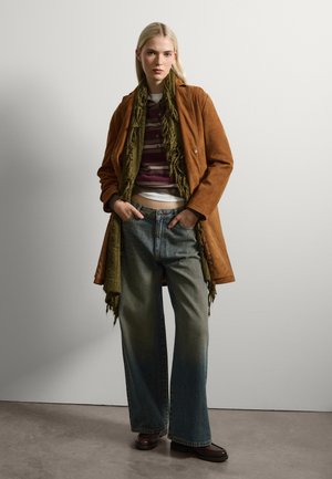 Manteau en daim marron, écharpe à franges vertes, pull rayé, jeans évasés délavés et chaussures marron. Textures lisses et palette de couleurs terreuses.