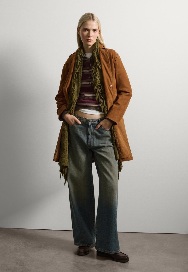 Cappotto in suede marrone, sciarpa frangiata verde, maglione a righe, jeans a gamba larga sbiaditi e scarpe marroni. Texture morbide e una palette di colori terrosi.