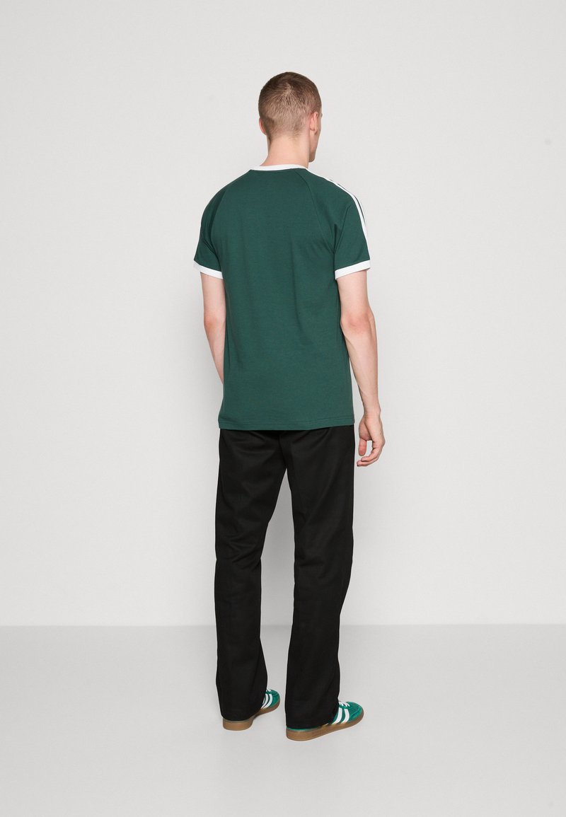 T-shirt en coton vert avec col blanc et bord des manches, porté avec un pantalon noir et des baskets vertes. Design sportif classique, vue de dos.