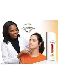 Le crème solaire Revitalift de L'Oréal dans un emballage blanc avec des accents rouges et jaunes, présenté à côté d'une femme portant un haut orange, recevant des conseils en soins de la peau.