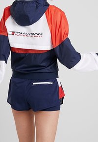 Osoba w granatowej, czerwonej i białej sportowej kurtce z kapturem Tommy oraz granatowych siatkowych szortach sportowych z białą zapinaną na zamek kieszenią z tyłu.