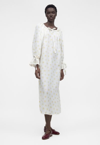 Γυναίκα που φοράει λευκό μίντι φόρεμα με κίτρινο λουλουδάτο σχέδιο, μακριά φουσκωτά μανίκια δεμένα στους καρπούς και μπορντό φλατ παπούτσια.