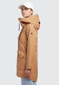 Braune wasserdichte Parka-Jacke mit Kapuze mit Kordelzug, Reißverschluss, zwei Fronttaschen und Druckknopf-Akzenten; getragen über hellblauen Jeans.
