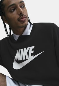 Sudadera negra con un gran logo blanco de Nike, presenta un cuello redondo y tejido texturizado. El modelo tiene el pelo trenzado y lleva una camisa blanca con cuello debajo.