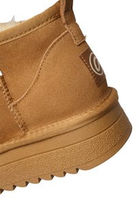 Close-up of tan suede ankle boot with textured rubber sole and stitched fabric pull tab featuring a circular logo.