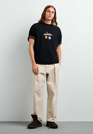 Giovane con i capelli fino alle spalle, che indossa una camicia nera a maniche corte con polsini arrotolati, pantaloni beige e stivali neri, in piedi contro un muro bianco.