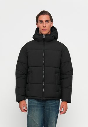 Ung mand iført en sort jakke med hætte og blå jeans, stående mod en simpel hvid baggrund.