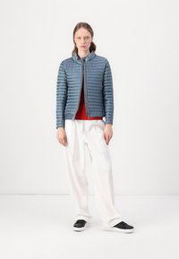 Pudret blå jakke med horisontal quiltet tekstur, lynlås lukning, båret over en rød top, parret med hvide bukser og sorte sneakers.