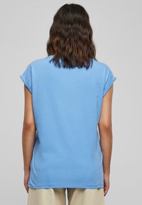 Kvinna med axellångt vågigt hår iklädd en ljusblå kortärmad skjorta och beige byxor, vänd bort från kameran mot en enkel grå bakgrund.