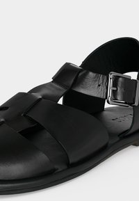 Μαύρο δερμάτινο σανδάλι με ανοιχτή μύτη, πλατιές λουρίδες, ρυθμιζόμενο ασημένιο αγκράφα και επίπεδη σόλα.