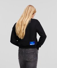 Sudadera negra de manga larga, con una etiqueta de parche azul en la parte posterior. Combinada con unos jeans gris oscuro, mostrando un look casual.